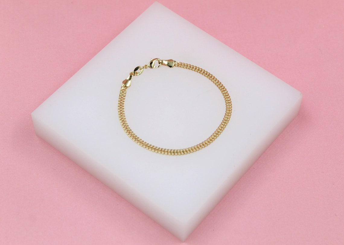 A delicate gold chain bracelet rests neatly on a white display block against a soft pink background.