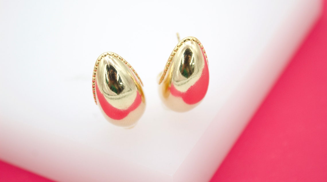 A pair of polished gold-plated drop earrings rests on a white display stand against a vibrant pink background.