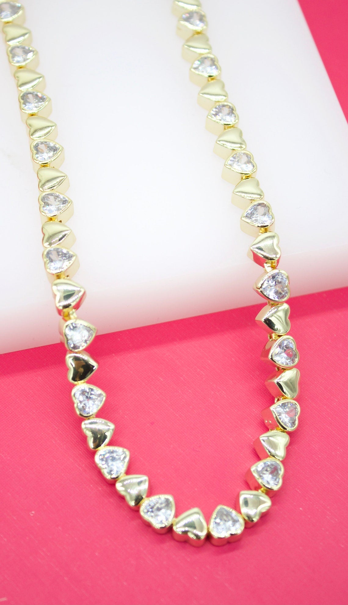 A gold heart-shaped tennis necklace with sparkling cubic zirconia stones rests against a pink and white background.