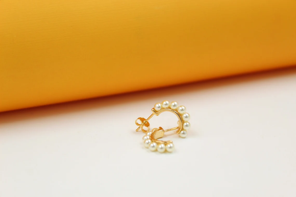 A pair of gold hoop earrings embellished with small white pearls rests on a light surface against an orange backdrop.