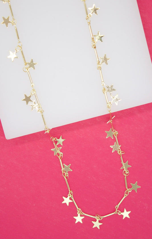 A delicate gold necklace features dangling star charms and small cylindrical bars arranged against a bright pink and white background.