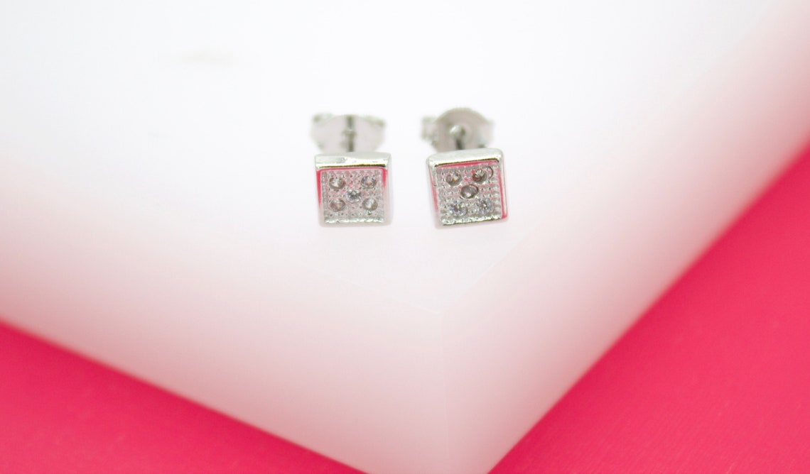 A pair of sparkling silver square stud earrings rests on a white display stand against a bright pink background.