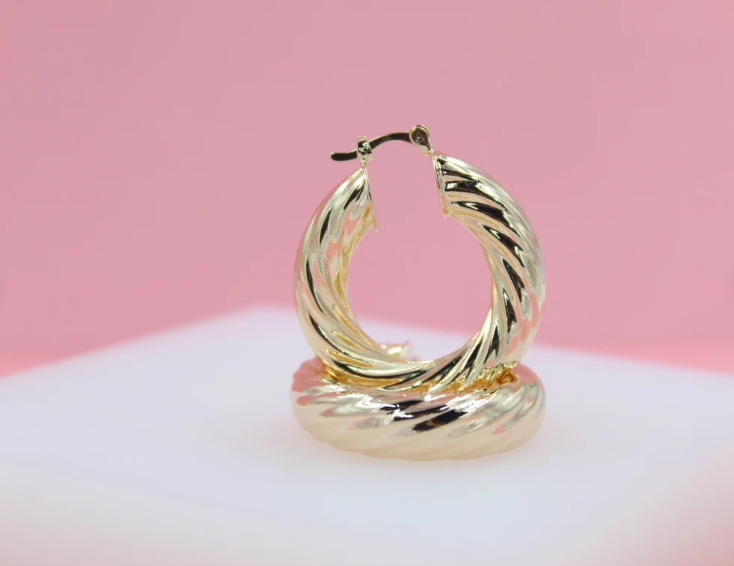 A pair of glossy gold twisted hoop earrings rests on a white platform against a soft pink background.
