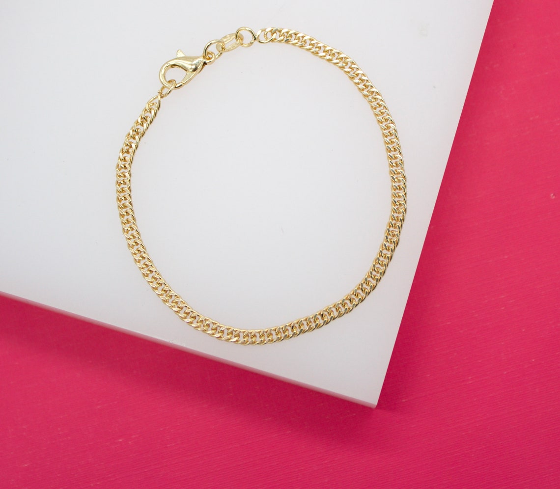 A delicate gold cuban link bracelet rests on a white display surface against a vibrant pink background.