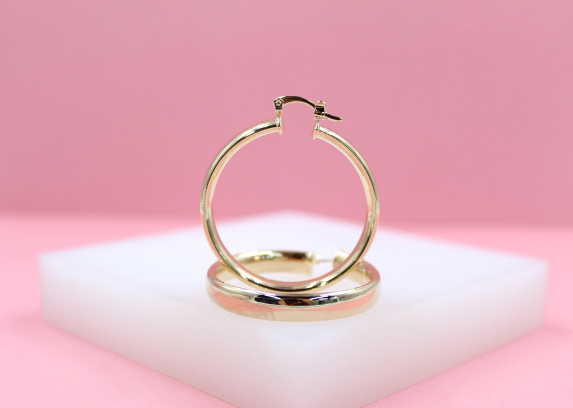 A pair of classic gold hoop earrings rests neatly on a white display block against a soft pink background.