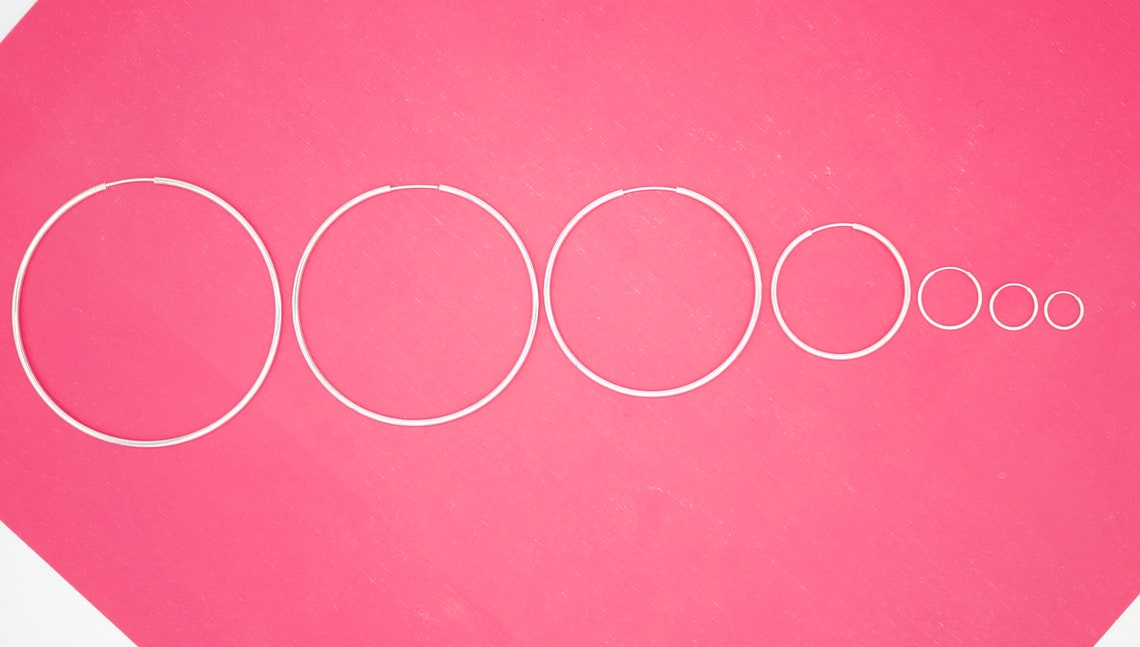 Seven silver hoop earrings in graduating sizes lie in a row against a vibrant pink background.