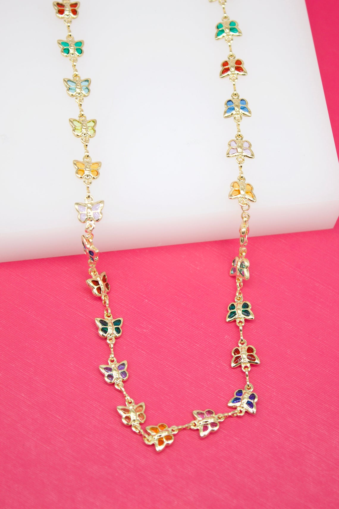 A gold butterfly necklace features colorful enamel accents in red, green, blue, and purple displayed against a bright pink background.