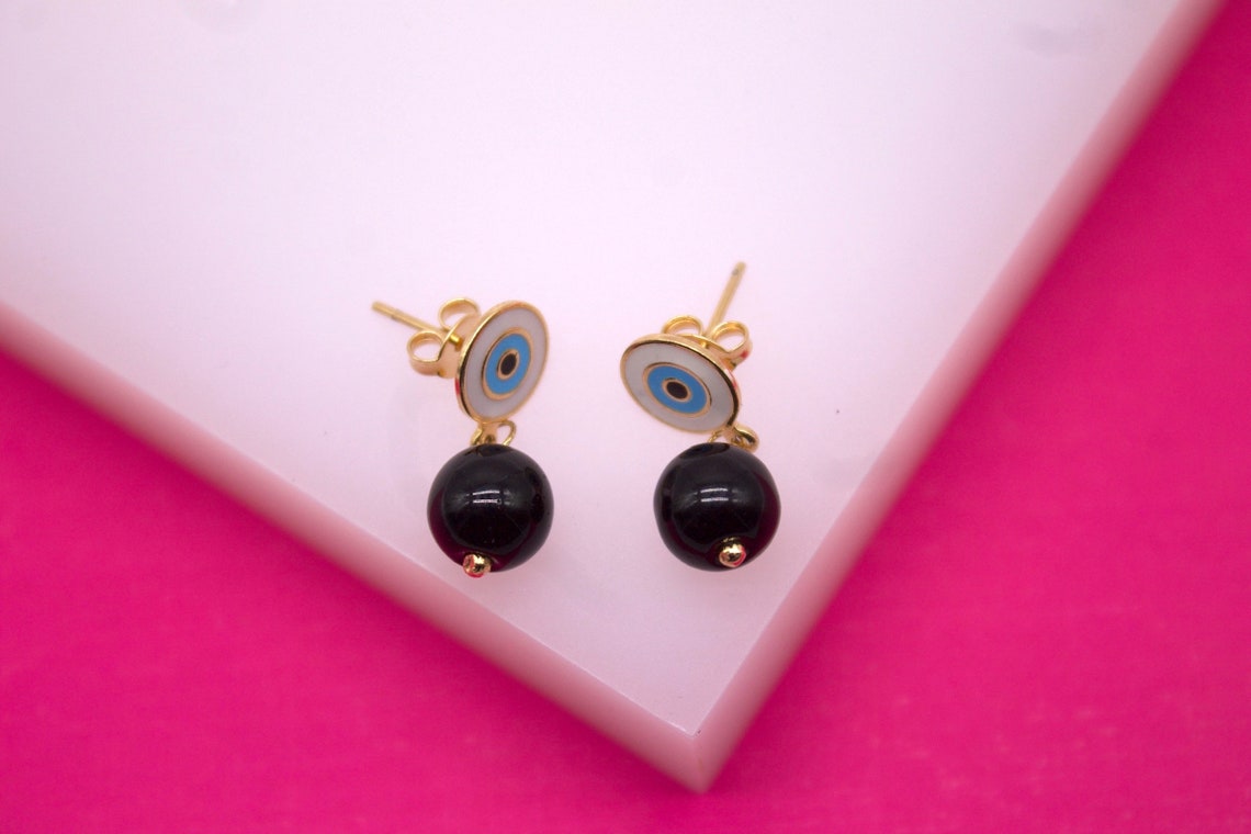Two gold evil eye stud earrings with dangling black beaded charms rest on a white display stand against a vibrant pink background.