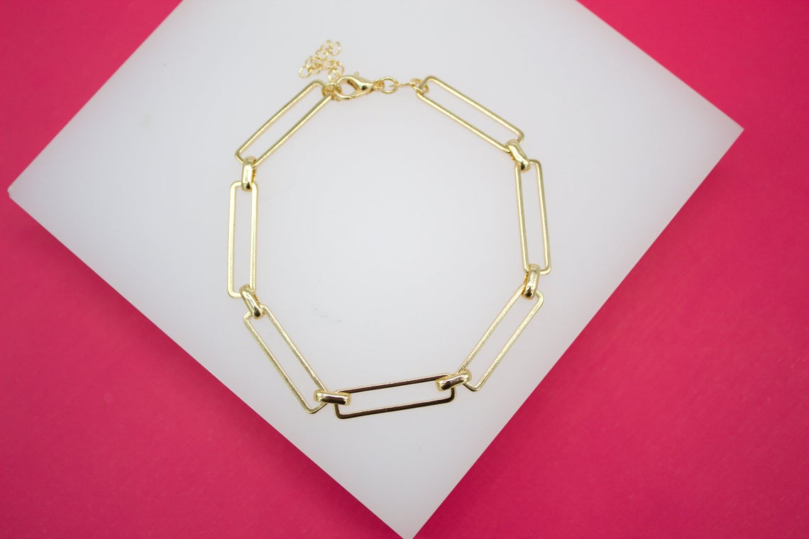 A stylish gold paperclip chain bracelet rests on a white display surface against a vibrant pink background.