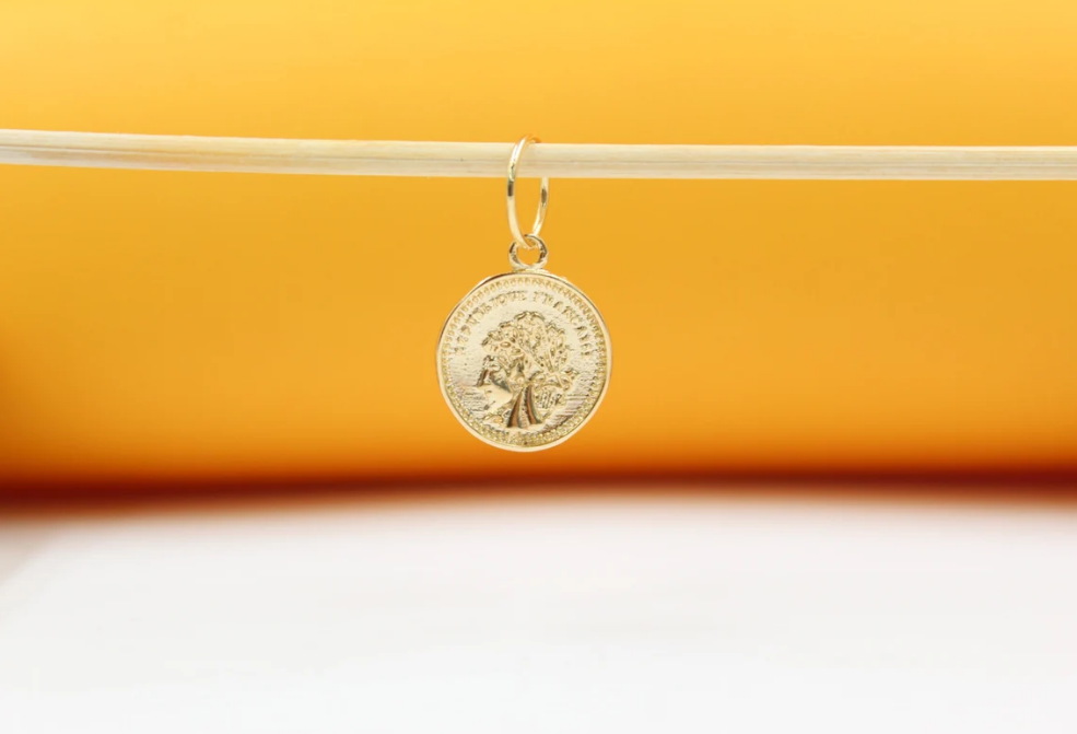 A delicate gold coin pendant hangs from a wooden stick against a warm yellow and white background.