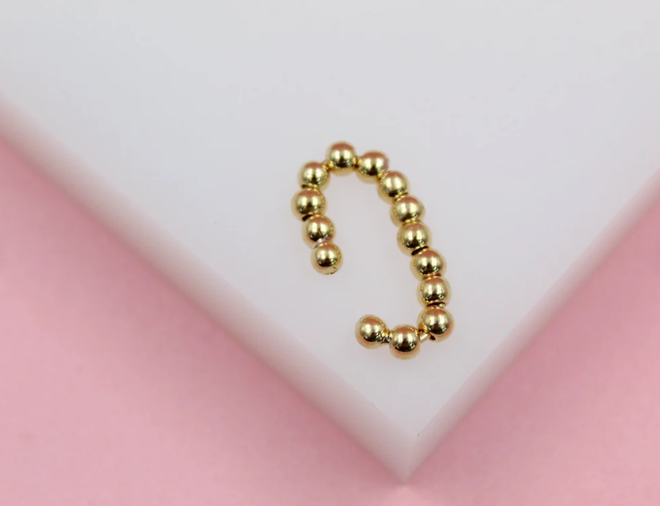 A sleek gold bead ear cuff rests on a white display against a soft pink background.