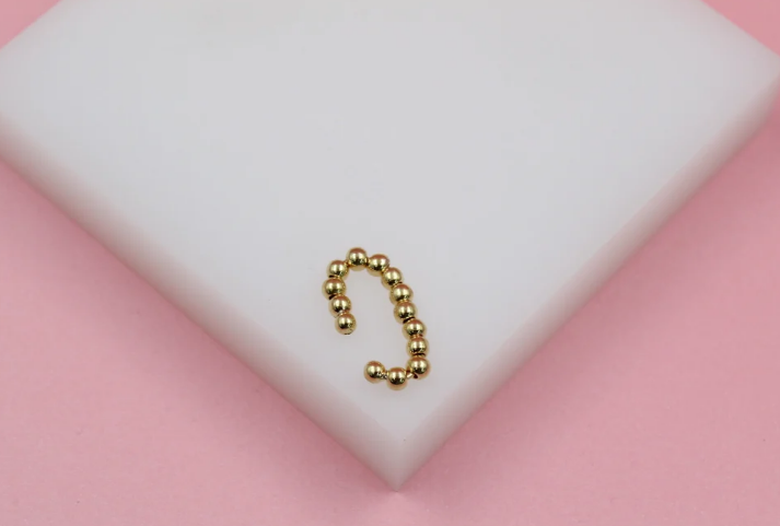 A gold beaded ear cuff rests on a white display block against a soft pink background.