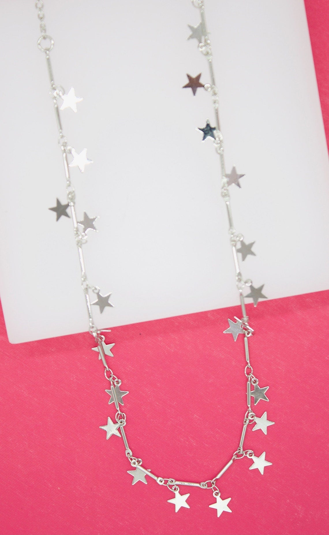 A delicate silver necklace features dangling star charms along its chain against a pink background.