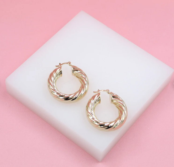 A pair of shiny gold twisted hoop earrings rests on a white block against a soft pink background.