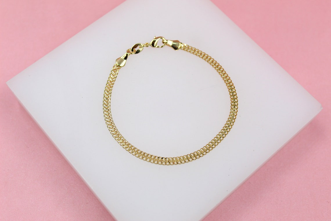 A delicate gold mesh chain bracelet rests on a white display stand against a soft pink background.