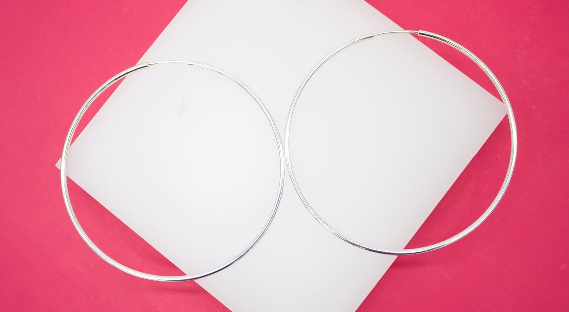 Two large sterling silver hoop earrings rest on a white display stand against a vibrant pink background.