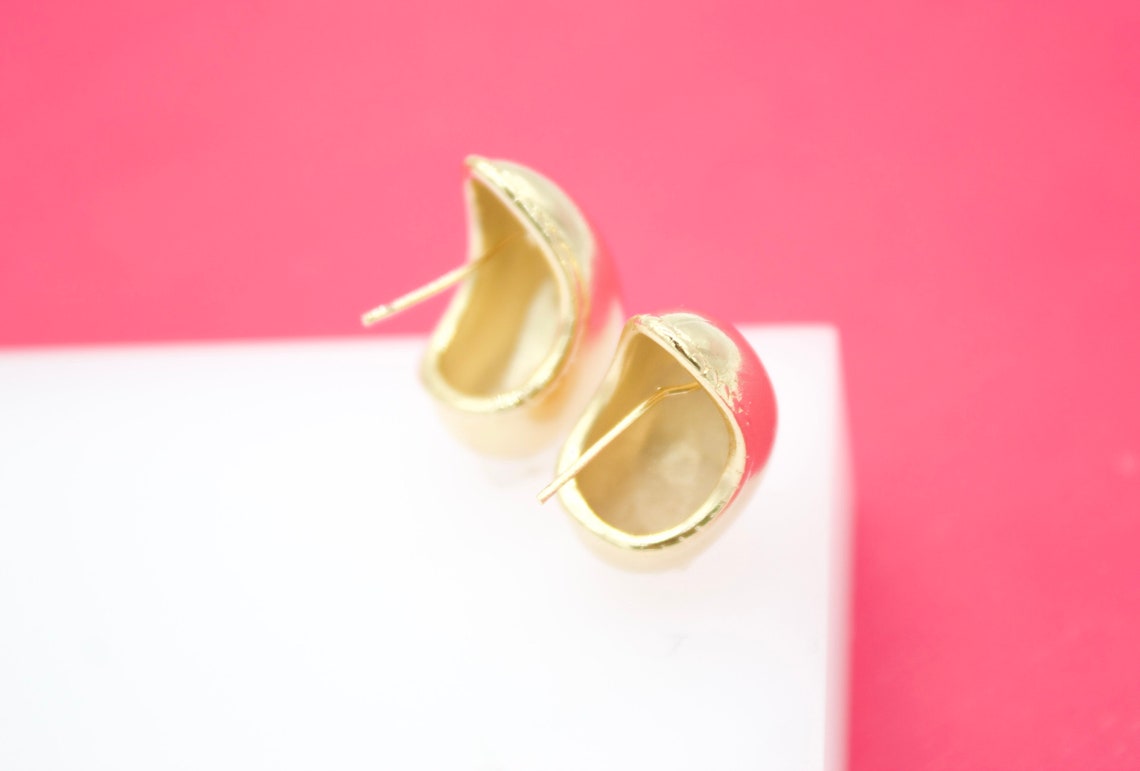 A pair of gold dome stud earrings rest on a white display against a vibrant pink background.
