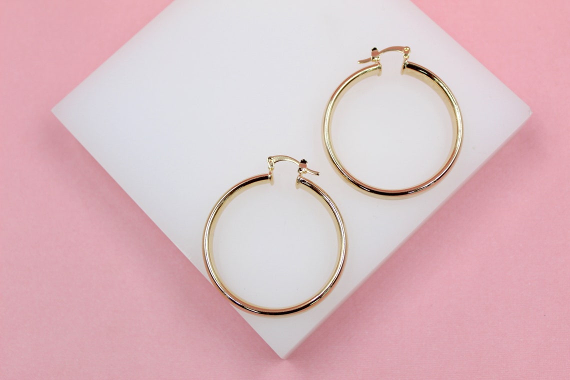 Two polished gold hoop earrings rest neatly on a white display block against a soft pink background.