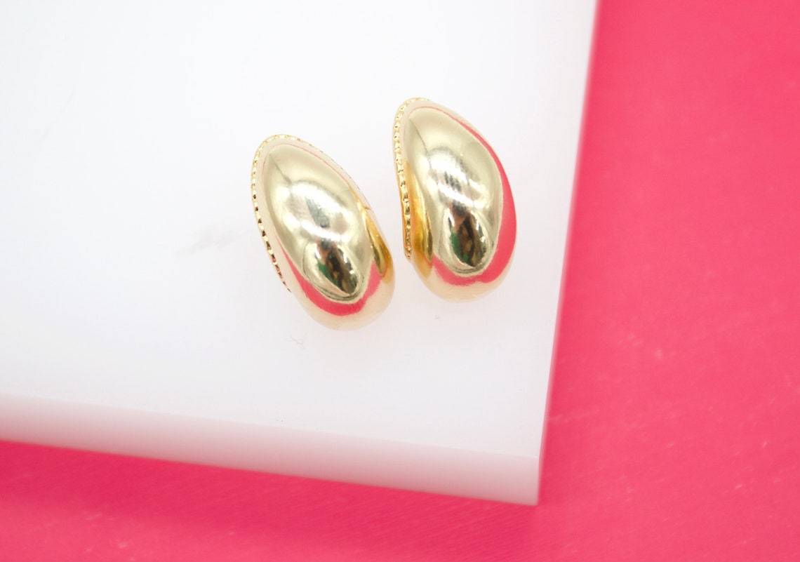 A pair of polished gold dome earrings rests on a white display block against a vibrant pink background.