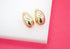 A pair of polished gold dome earrings rests on a white display block against a vibrant pink background.