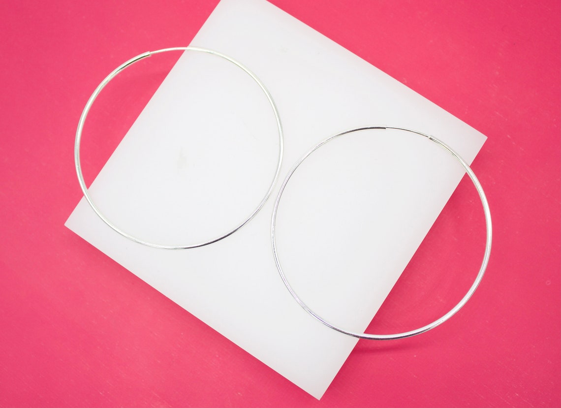 Two large silver hoop earrings rest on a white square display against a vibrant pink background.