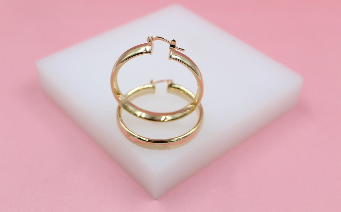 A pair of polished gold hoop earrings rests upright on a white display block against a soft pink background.