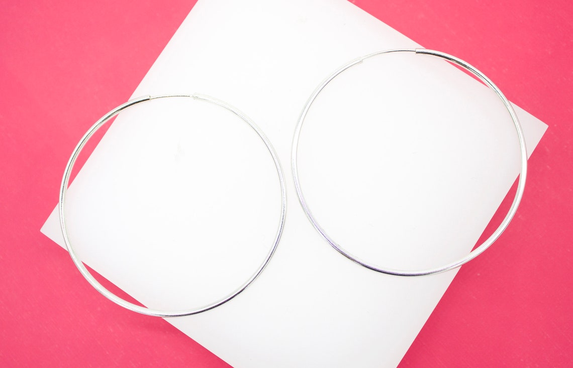 Two large silver hoop earrings rest on a white display surface against a vibrant pink background.