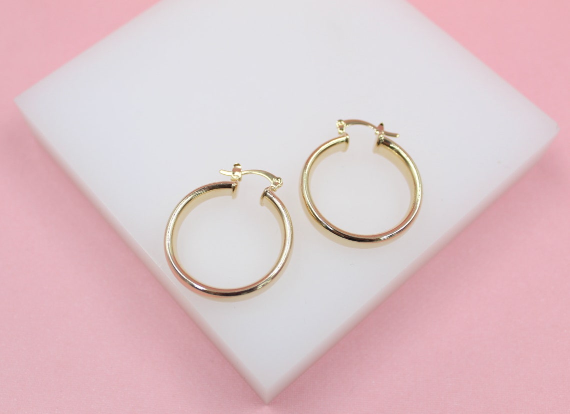 A pair of shiny gold hoop earrings rests on a white display block against a soft pink background.