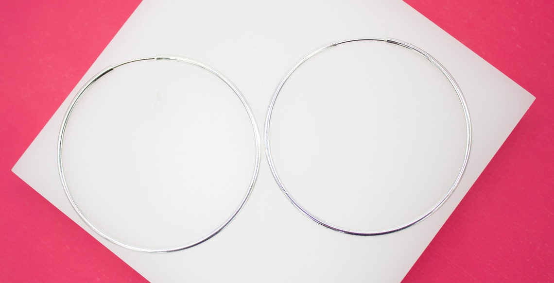 A pair of sleek silver hoop earrings rests neatly on a white display stand against a vibrant pink background.
