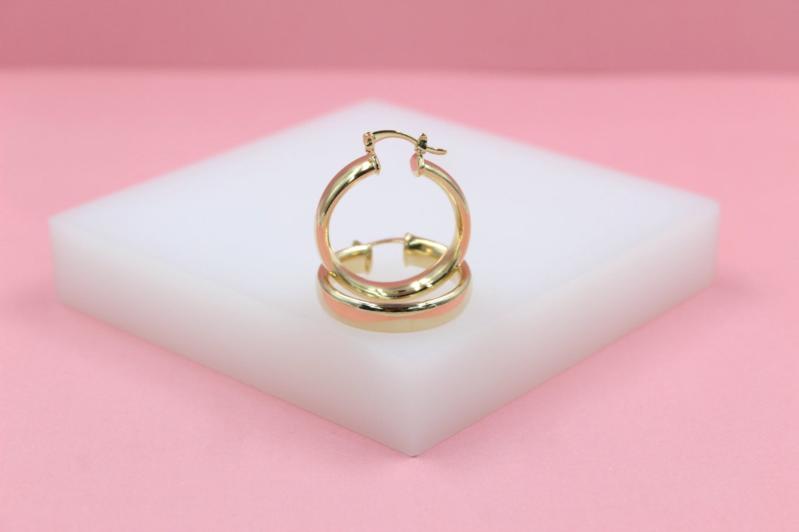 A pair of glossy gold hoop earrings rests upright on a white platform against a pink background.