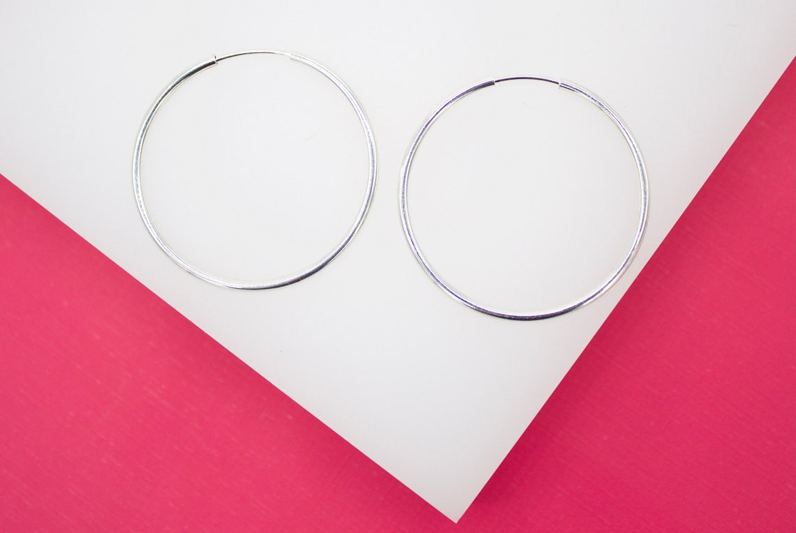 Two large silver hoop earrings lie flat against a white display surface bordered by bright pink.