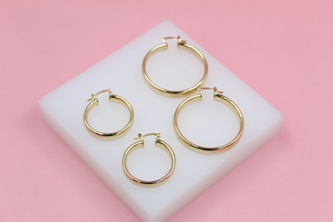 Four glossy gold hoop earrings in varying sizes rest on a white display block against a soft pink background.