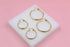 Four glossy gold hoop earrings in varying sizes rest on a white display block against a soft pink background.