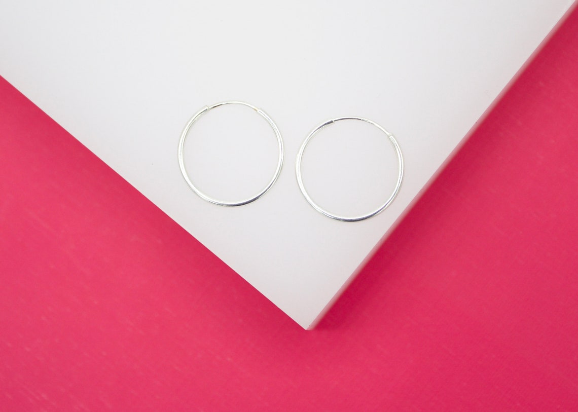 A pair of simple silver hoop earrings rests neatly on a white display surface against a vibrant pink background.