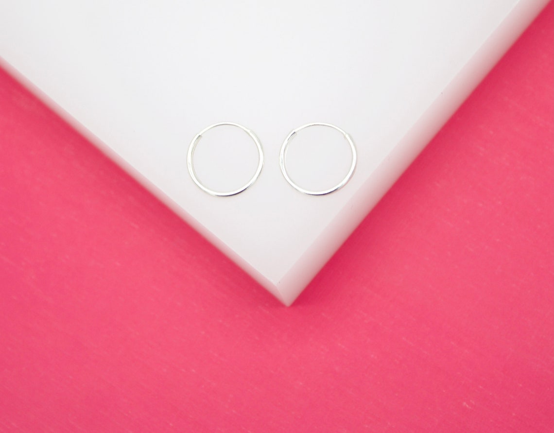 A pair of minimalist sterling silver hoop earrings rests on a white platform against a vibrant pink background.