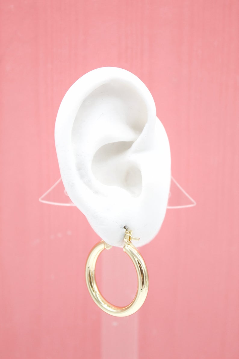 A thick gold hoop earring hangs from a white model ear against a pink background.