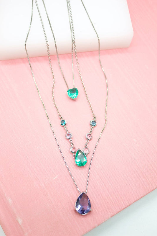 Three delicate silver necklaces with colorful crystal pendants rest on a pink surface.