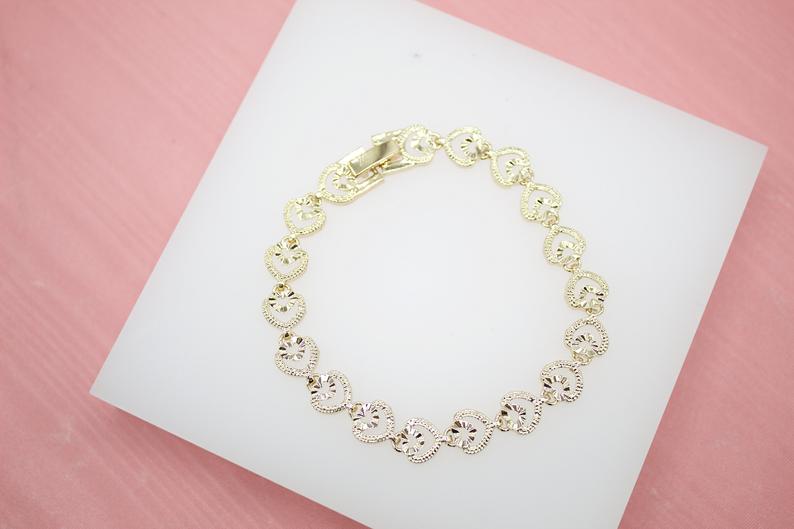 A delicate gold heart link bracelet rests on a white display surface against a pink background.