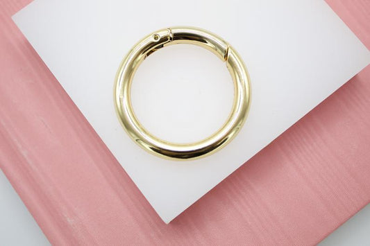 A shiny gold spring gate ring rests centrally on a white square surface against a soft pink background.