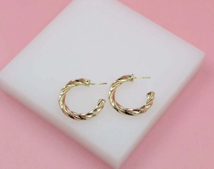A pair of polished gold twist hoop earrings rests on a white block against a soft pink background.