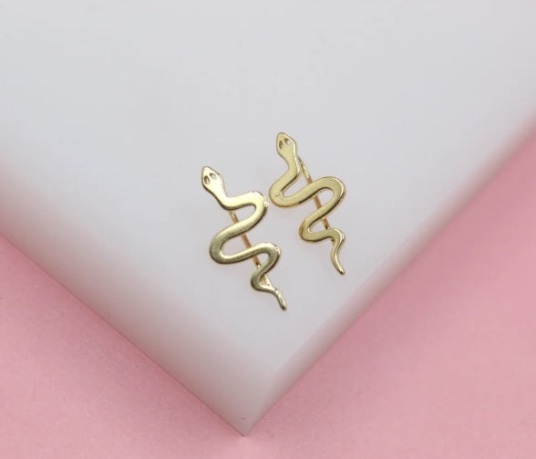Two gold snake crawler earrings rest on a white display against a pink background.