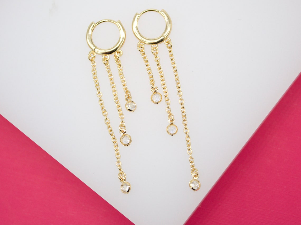 A pair of gold huggie hoop earrings featuring three dangling chains set with sparkling cubic zirconia stones rests against a white and pink backdrop.