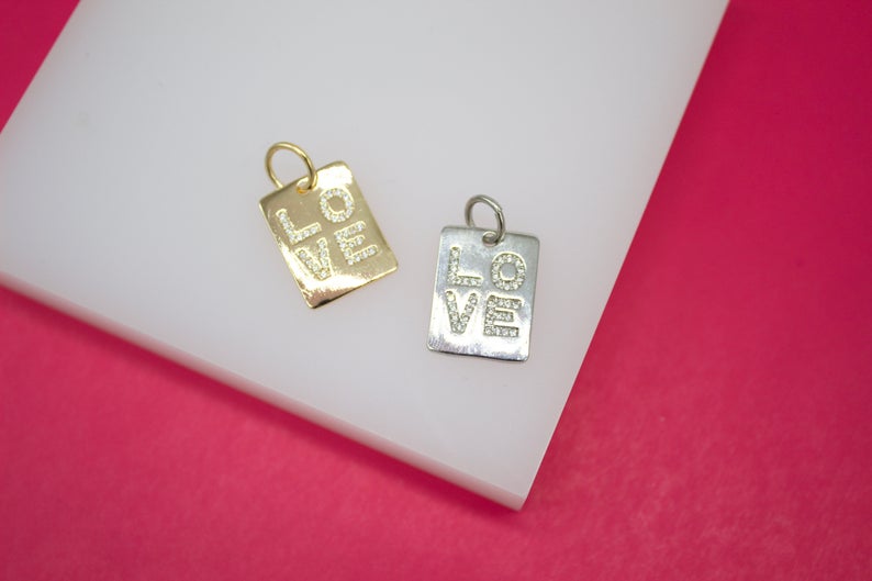 Two rectangular jewelry charms, one gold and one silver, feature pavé-set diamond letters spelling out the word "LOVE" against a white and pink background.