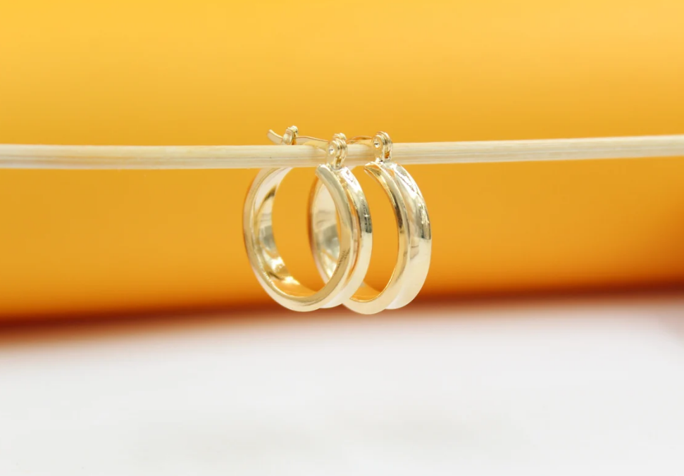 Two polished gold hoop earrings hang delicately from a thin horizontal stick against a warm yellow and white background.