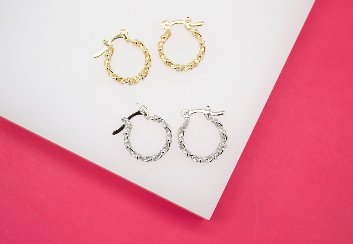 Two pairs of small twisted huggie hoop earrings, one in gold and one in silver, rest on a white display against a pink background.