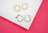 Two pairs of small twisted huggie hoop earrings, one in gold and one in silver, rest on a white display against a pink background.