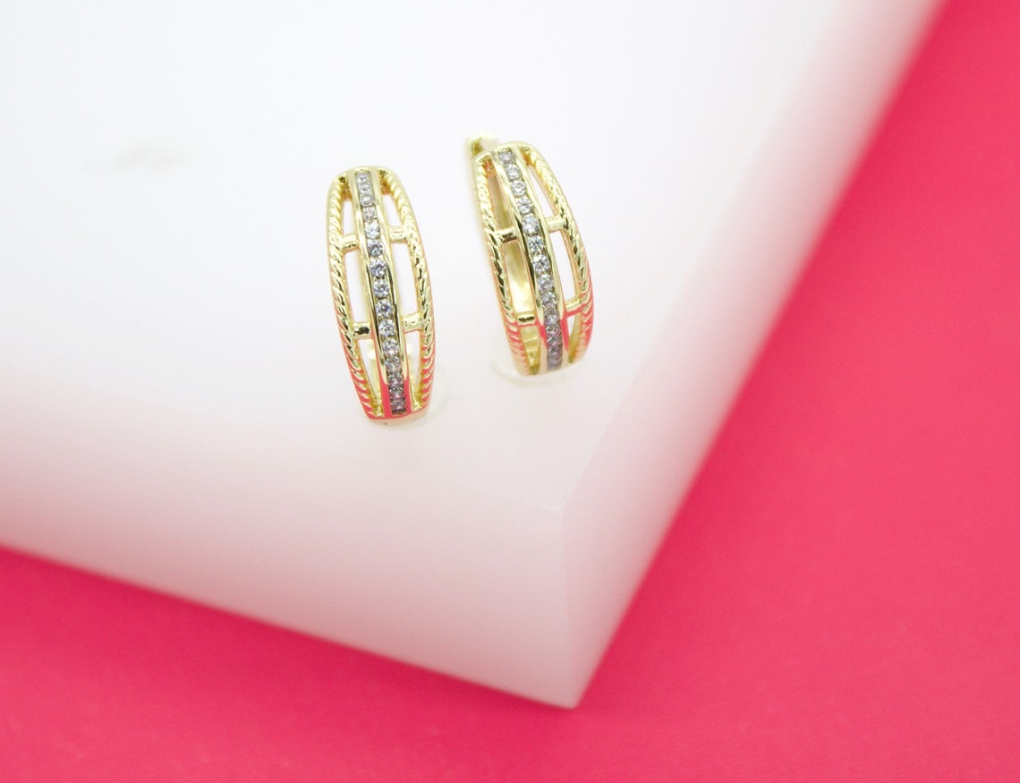 A pair of elegant gold hoop earrings featuring sparkling pavé diamond centers rest upright on a white display block against a vibrant pink background.