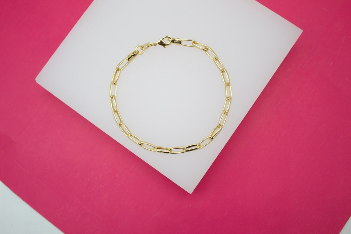 A gleaming gold paperclip chain bracelet rests on a white display against a vibrant pink background.