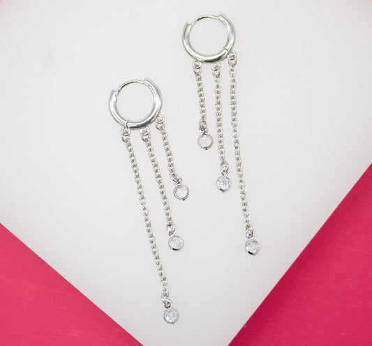 A pair of silver hoop earrings features three delicate dangling chains tipped with sparkling crystal stones, displayed against a pink and white background.