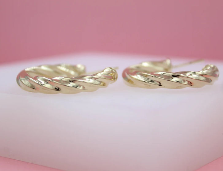 Two glossy, twisted gold hoop earrings rest side-by-side on a white surface against a soft pink background.
